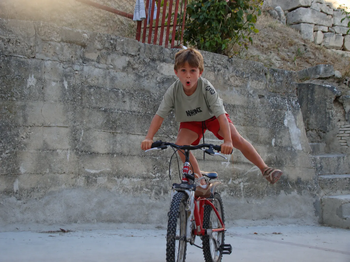Niño con camiseta NONI verde oliva haciendo un truco sobre la bici al atardecer