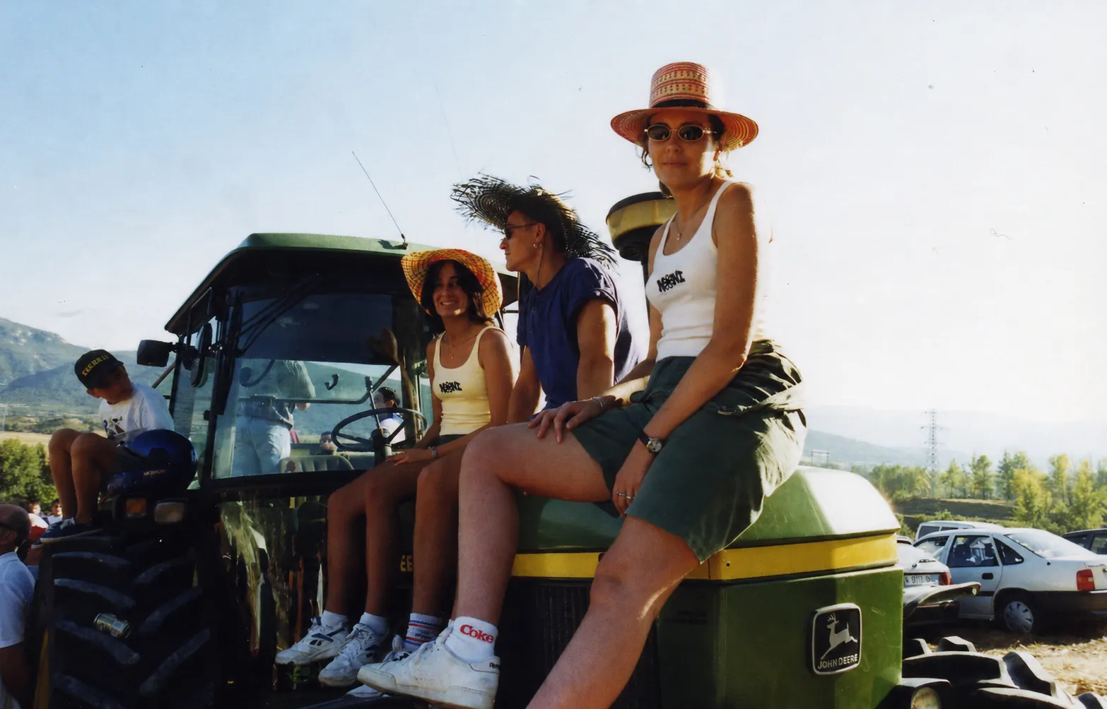 Tres personas subidas a un tractor John Deere en el Valle de Tobalina; una joven con camiseta de tirantes NONI mira a cámara bajo la luz dorada del verano.